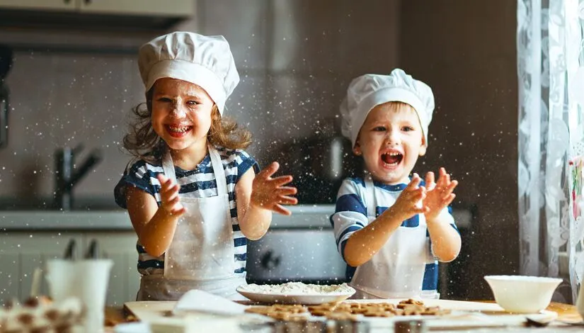 Young brother and sister baking cookies. Wearing aprons and chefs hats and clapping and laughing.