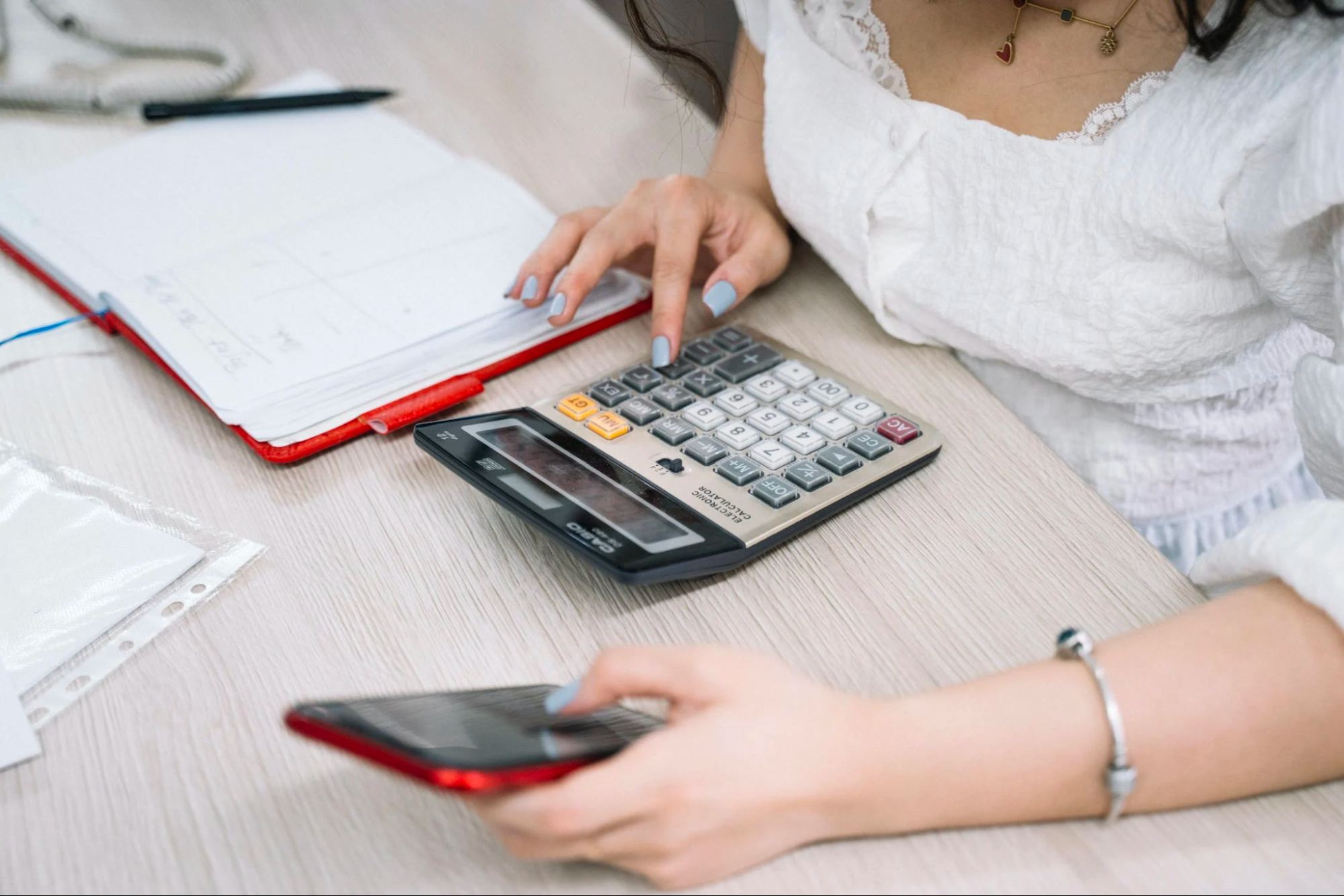 Woman using phone and calculator and datebook at desk.