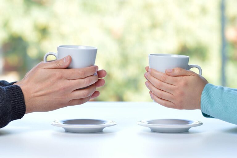 spouses sitting across from one another having coffee. hands wrapped around coffee mugs. bright day.