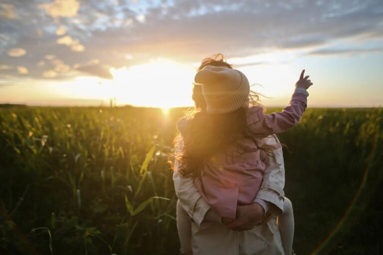 mother holding child in pink coat standing in a field. Bright sunset.