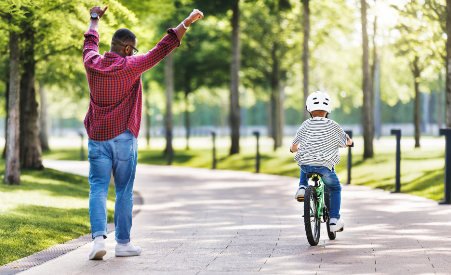 Dad walking alongside his child who is riding bike independently. Dad's arms are extended in the air in excitement. Park setting.
