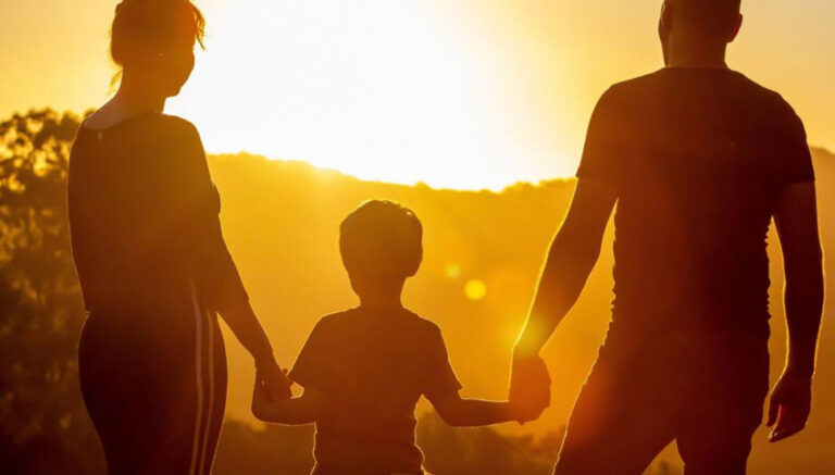 Young male child holding hands with his parents, silhouetted against bright evening sunset.