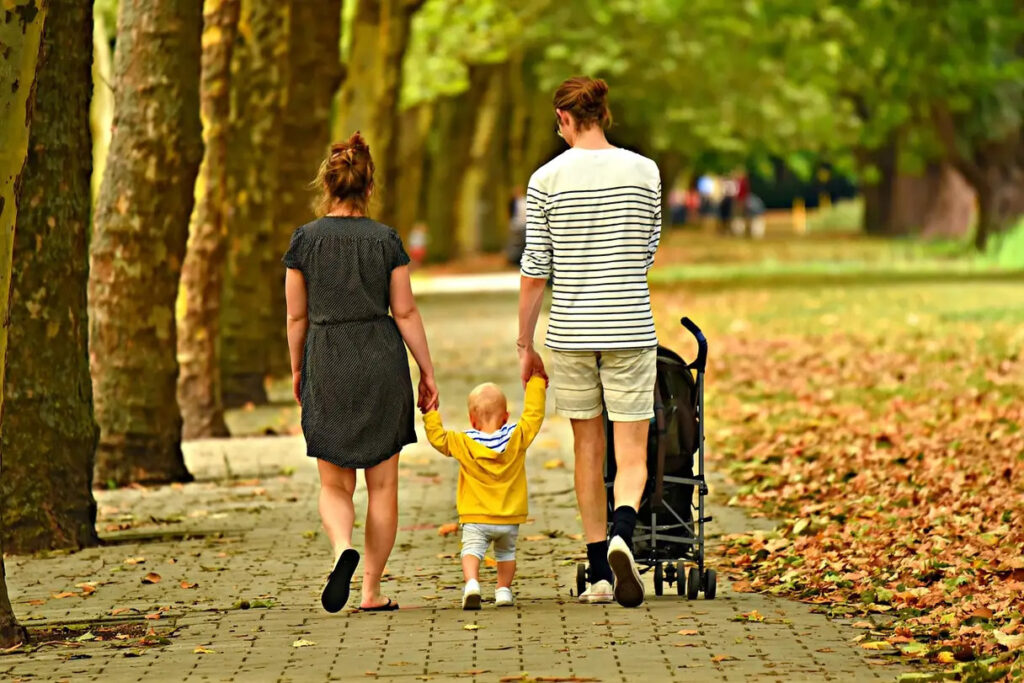 mom and dad holding hands with toddler, walking away down a treelined path on fall day.