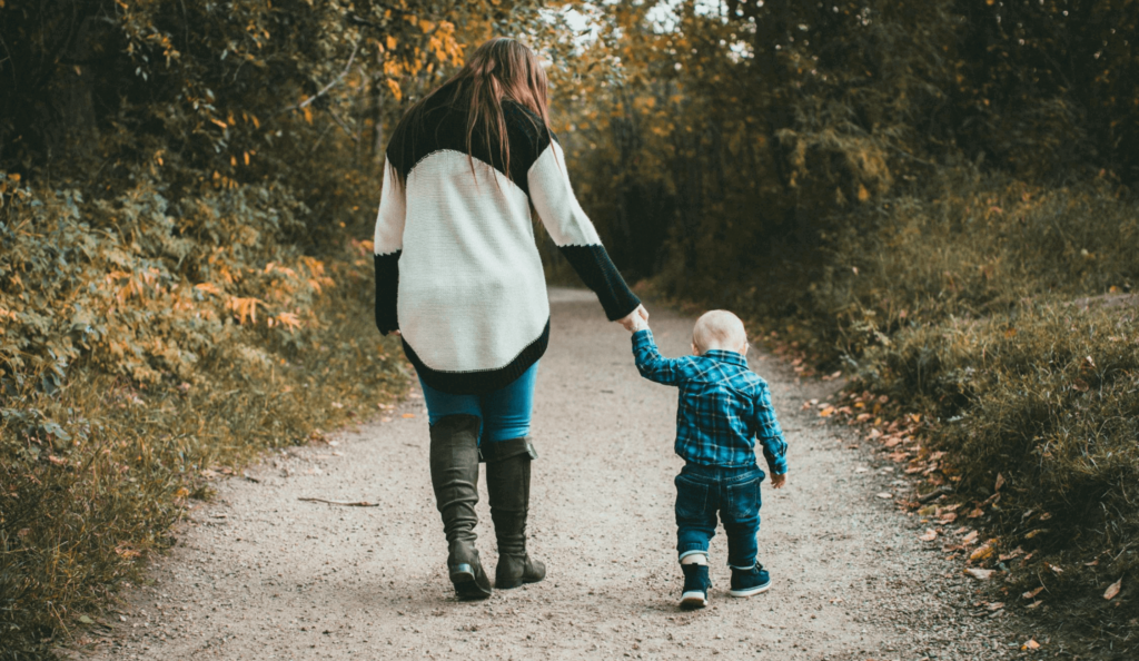 Mom in sweater and boots and baby in flannel shirt walking down a gravel path in fall.