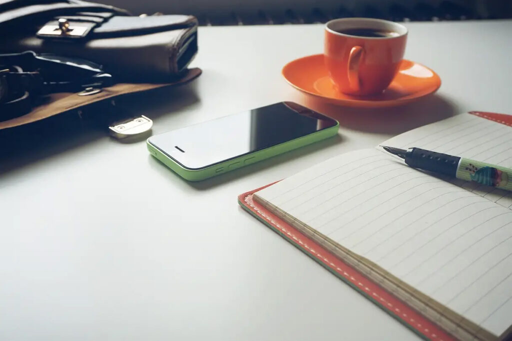 Noteboook with pen, phone and orange cup of coffee.