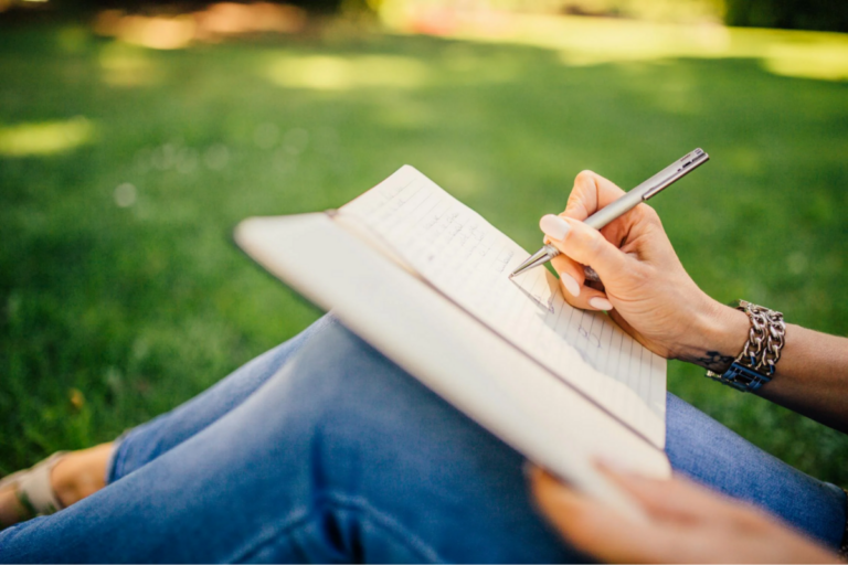 Woman's knees with notebook propped on them and she is writing in it. bright green grass background.