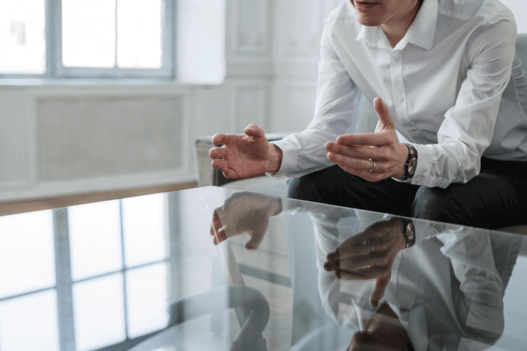 Man in white shirt sitting on couch, leaning over glass coffee table, gesturing stressfully.