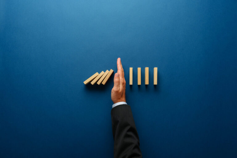 Blue background, mans arm and hand, wearing suit. on left side of his hand are 4 wooden blocks that are falling and on the right are 4 wooden blocks that are not falling.