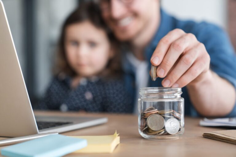 Dad's hand dropping coins in a jar. Child and dad blurry in the background.