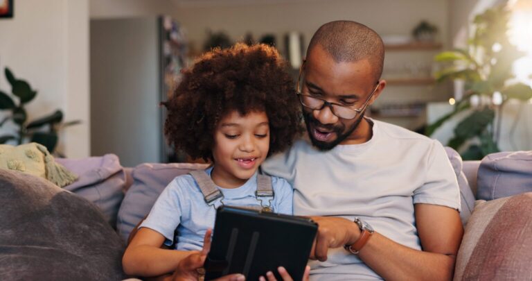 Dad and son sitting on the couch looking at an ipad. Smiling. Home environment with plantsin background.