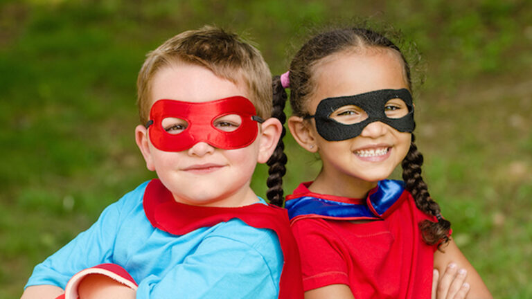 two young kids wearing capes, masks and with arms folded in front of them. Smiling.