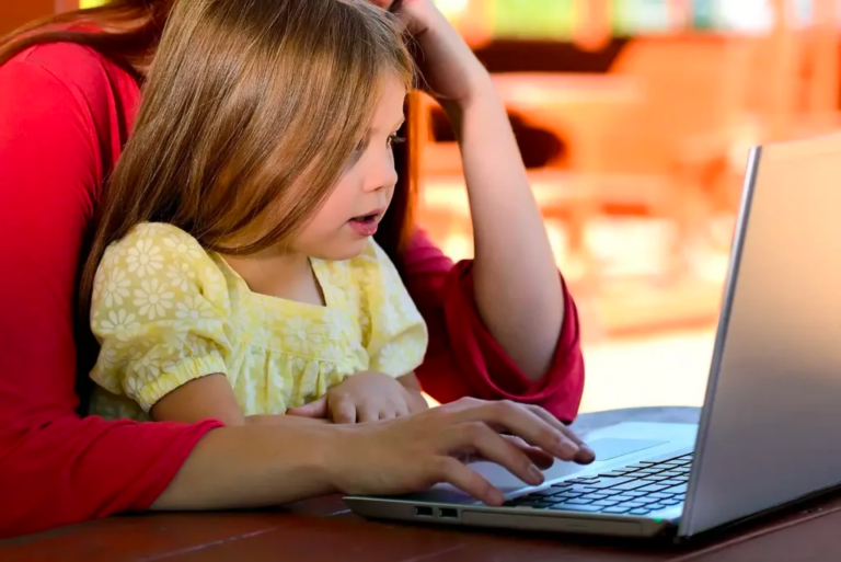 Young girl in yellow blouse, sitting on mom's lap as they look at a laptop. Child is very interested.