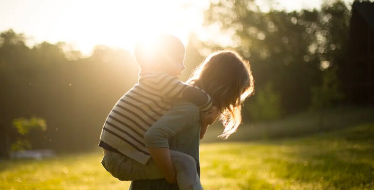 Sister carrying her brother piggyback. Green grass on sunny evening