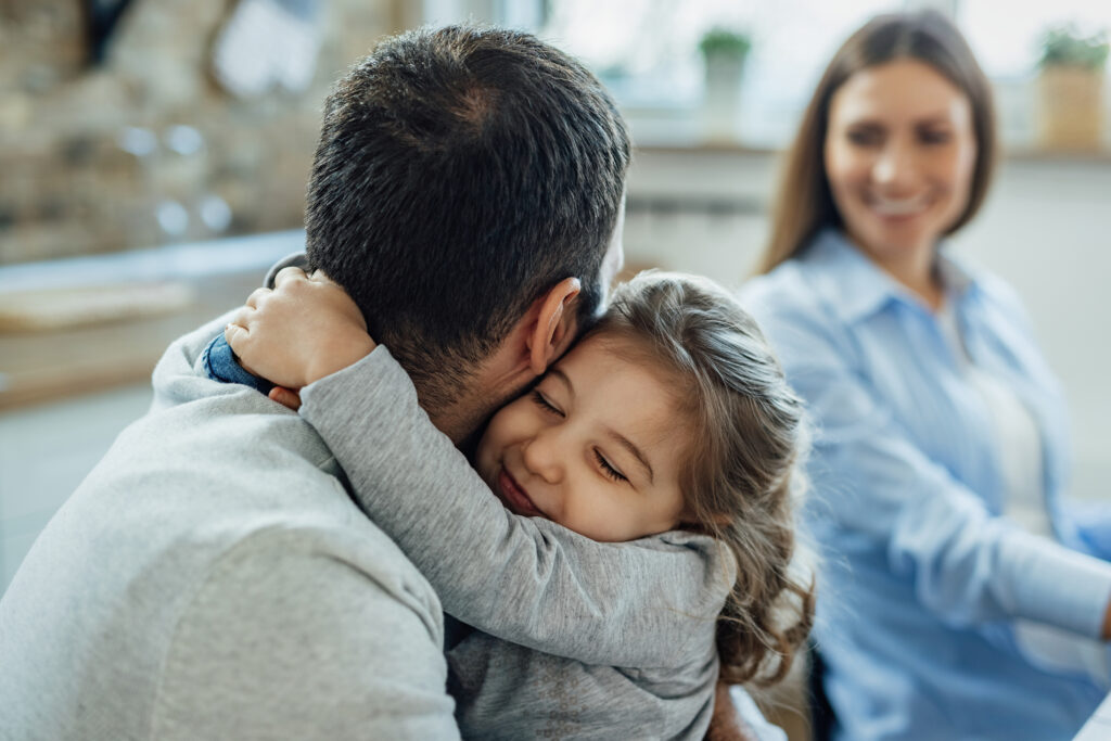 affectionate little girl with eyes closed embracing her father home mother is background