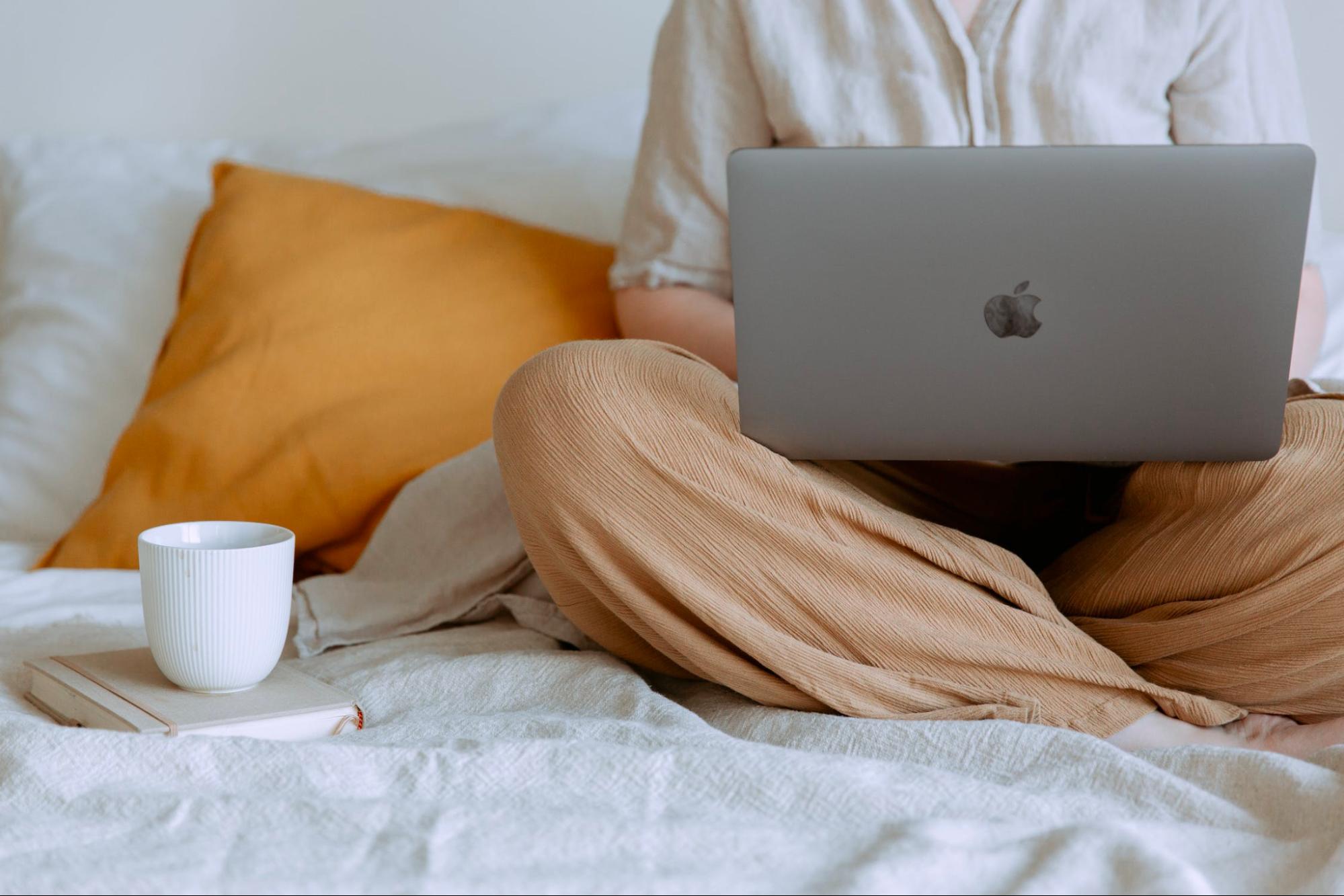 Image of a person typing at their computer on their bed, with a cup of coffee nearby.