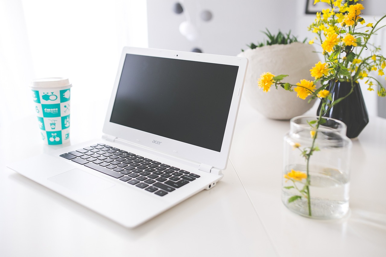 A laptop is placed in between a coffee cup and a jar containing yellow flowers.