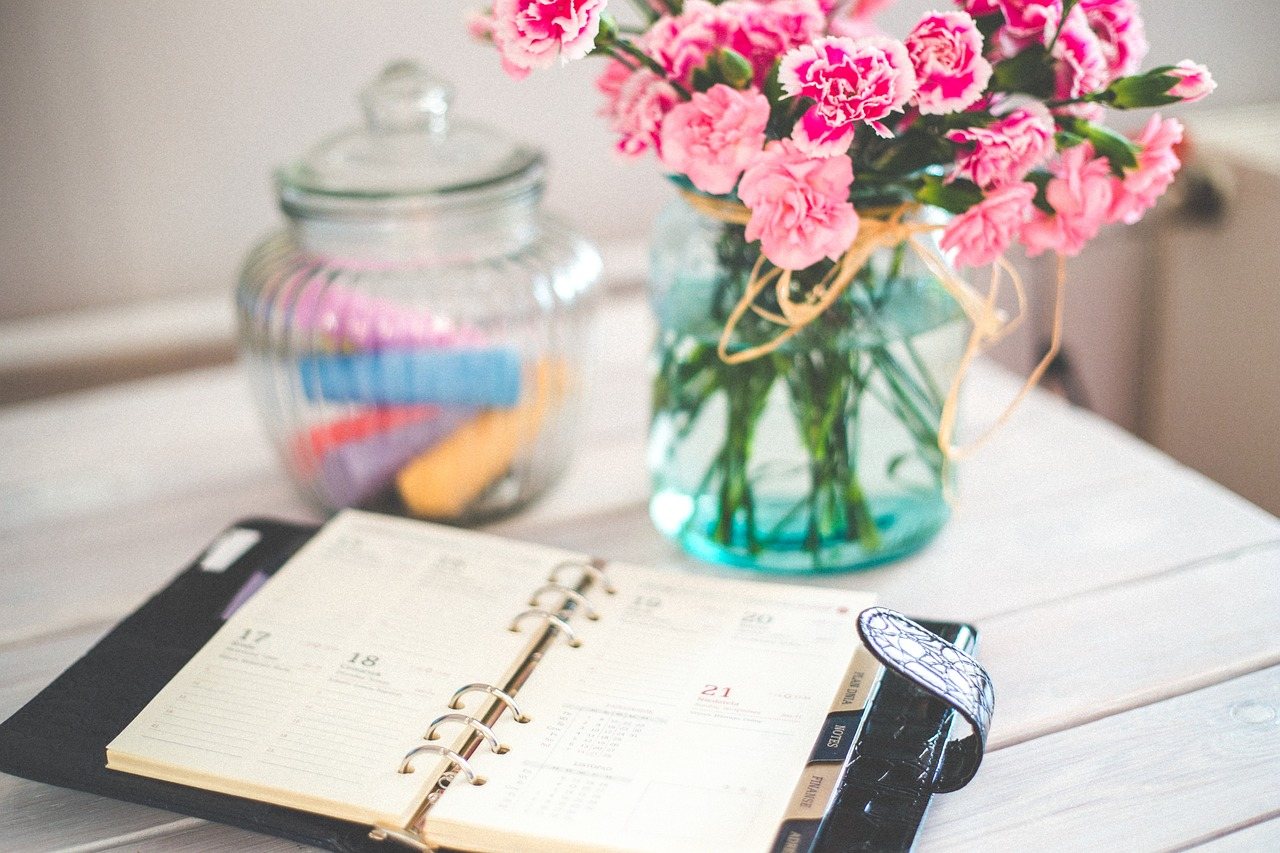 A planner lies on a table, along with a vase of flowers and a jar.