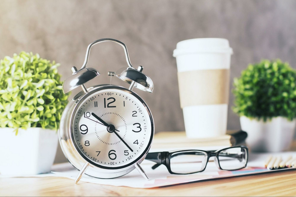 This is an image of an alarm clock, a pair of glasses, two plants, and a cup of coffee on a table.