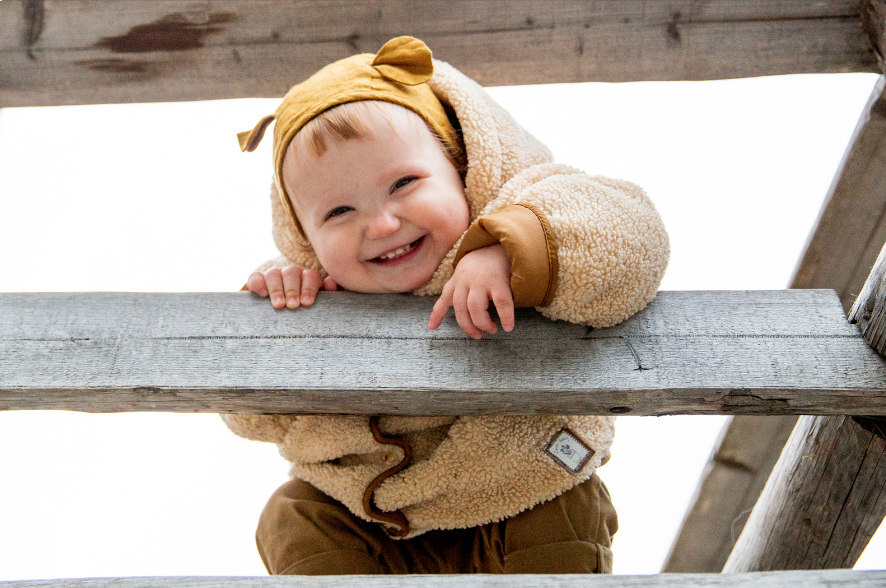 This is an image of a toddler leaning on a wooden fence rail and smiling.