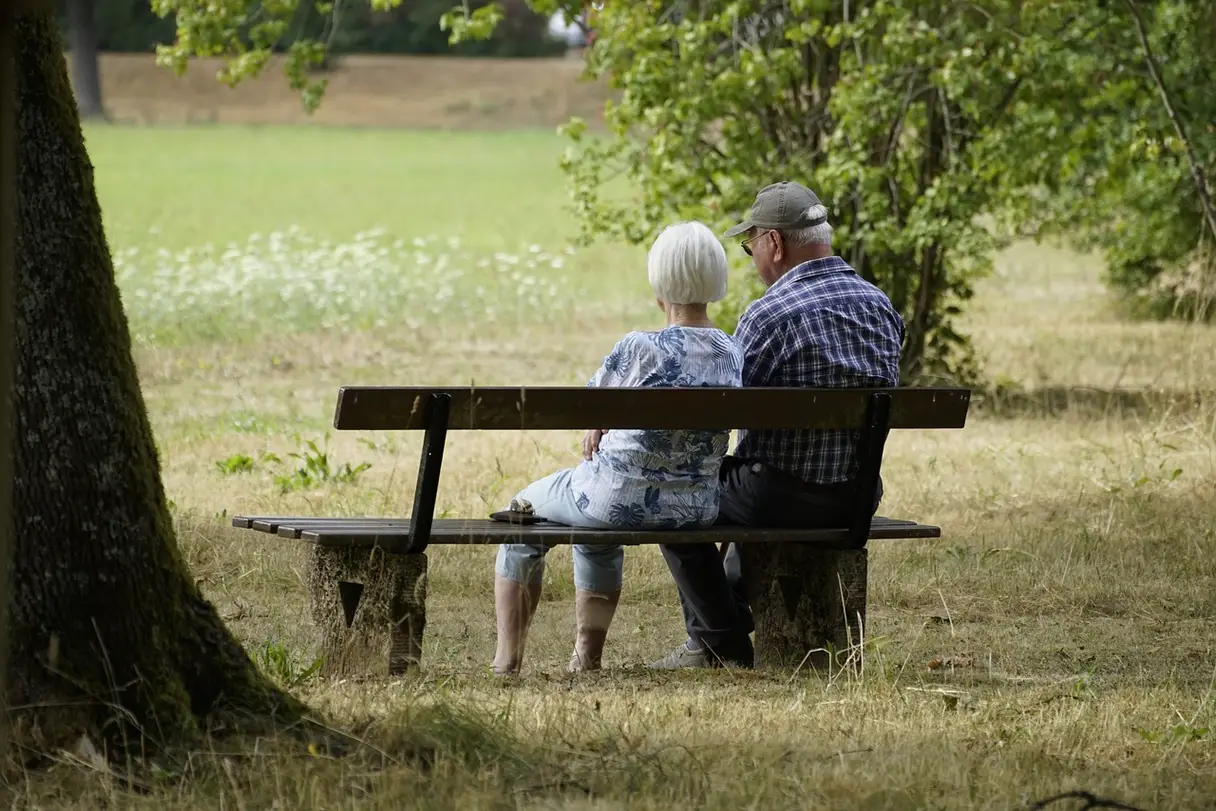 This is an image of two elderly individuals sitting together on a bench outside.